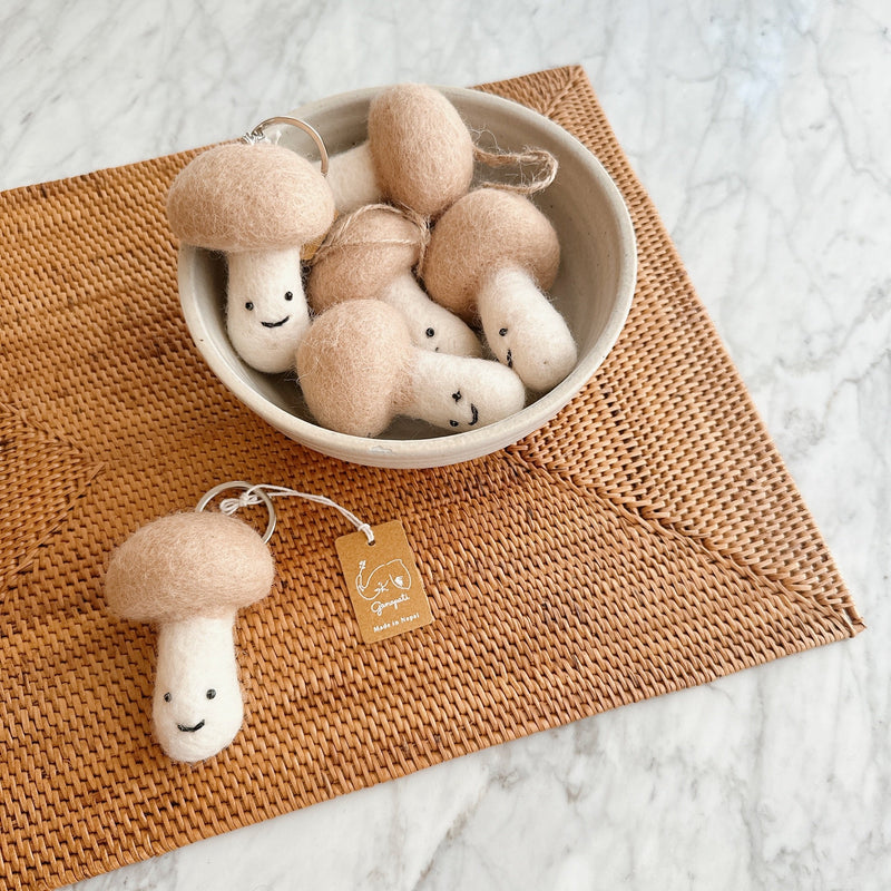 Smiley Beige Mushroom felt ornaments are displayed, with one resting on a rustic woven rattan placemat and others nestled in a gray bowl, against a marble backdrop. Each charming handmade felt ornament features a rounded beige cap and a white stem, complete with a small, cheerful smiling face. A silver ring and "Ganapati, Made in Nepal" tag enhance this delightful, handcrafted display.