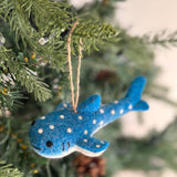 A Smiley Whale Shark felt Christmas ornament hangs by a brown jute string on a vibrant green Christmas tree. This cheerful felt whale shark ornament is blue, featuring a soft white underbelly, playful white polka dots, and a distinctive smiley design. The warm lights of the tree enhance its festive appearance.