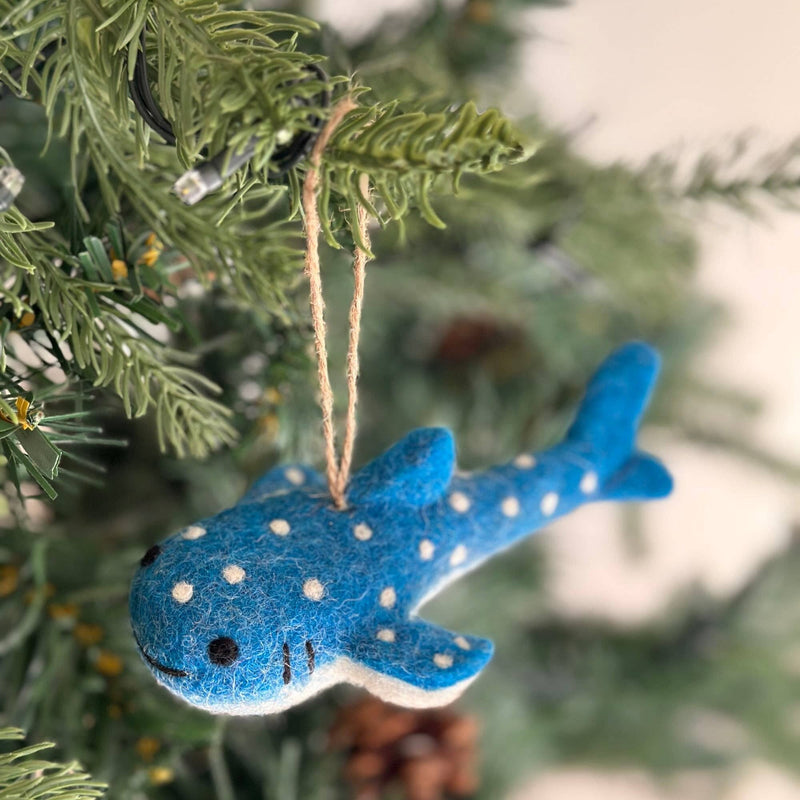 A Smiley Whale Shark felt Christmas ornament hangs by a brown jute string on a vibrant green Christmas tree. This cheerful felt whale shark ornament is blue, featuring a soft white underbelly, playful white polka dots, and a distinctive smiley design. The warm lights of the tree enhance its festive appearance.