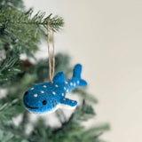 A smiley whale shark felt ornament hangs on a Christmas tree branch against a soft white background. This blue felt whale shark ornament features white spots and stitched smile and eyes, adding a cheerful, whimsical touch. A jute string is attached for easy hanging.