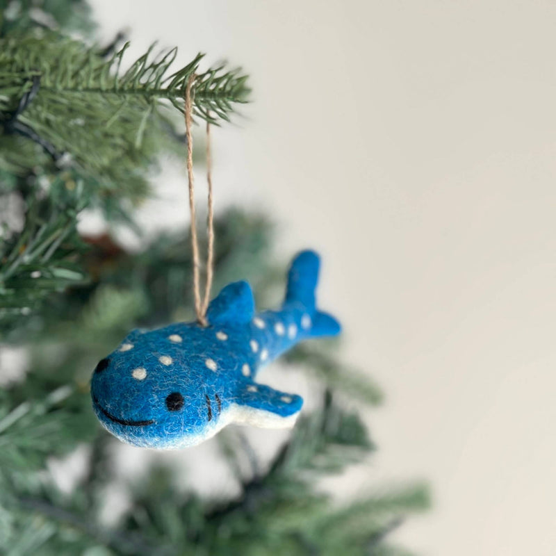 A smiley whale shark felt ornament hangs on a Christmas tree branch against a soft white background. This blue felt whale shark ornament features white spots and stitched smile and eyes, adding a cheerful, whimsical touch. A jute string is attached for easy hanging.