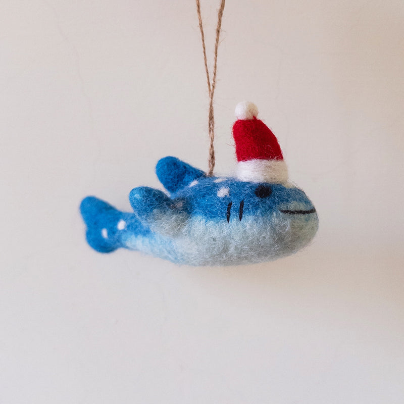 A playful Smiley Whale Shark felt ornament hangs suspended by a jute string against a plain, light background. This handcrafted felt ornament features a gradient from dark to light blue, adorned with white spots and black stitch marks suggesting eyes and a smile.
