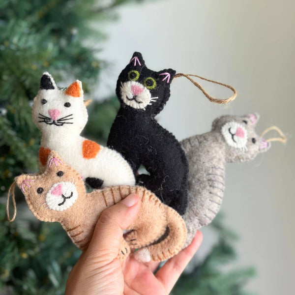 A female hand holds four charming wool felt stitched cat ornaments against a soft-focus Christmas tree background. One light brown cat ornament lies horizontally in the palm, while a white and orange, a black, and a grey cat ornament are stacked vertically. Each unique Christmas tree ornament features delicate stitched eyes, nose, and whiskers, suspended by a natural jute string.