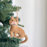 A handcrafted stitched cat felt ornament hangs from a verdant Christmas tree branch, exuding cozy, festive charm. This tan wool felt cat ornament features darker tan stitched stripes, a pink nose, and white whiskers. A jute string at the top allows for easy hanging, enhancing seasonal cheer.