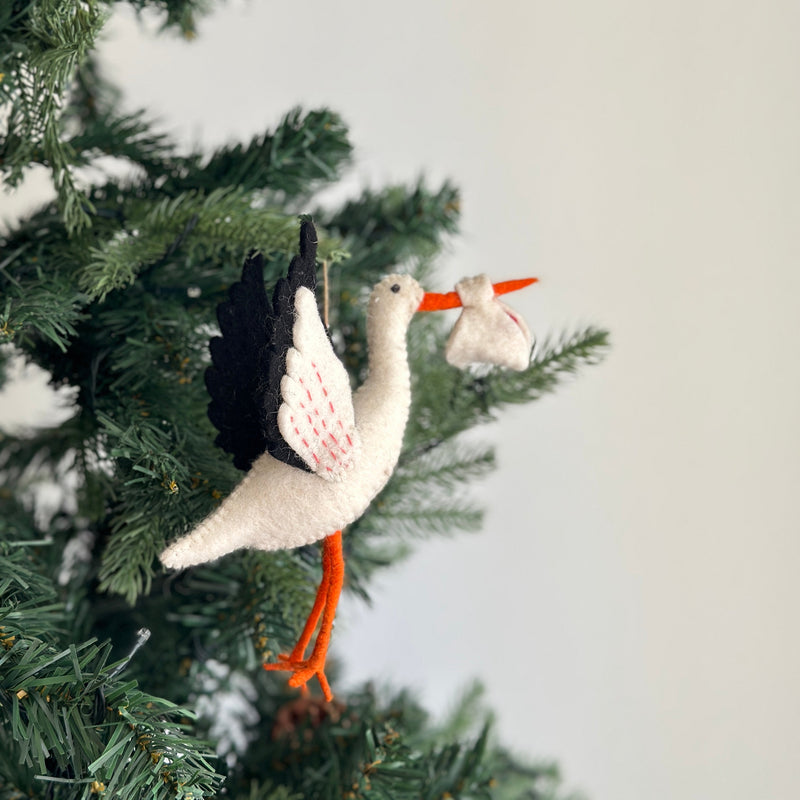 A felt stork ornament is suspended from a green pine branch, appearing in mid-flight, against a blurred backdrop. The hand-felted stork has a beige body, black and white wings detailed with red stitching, an orange beak, and orange legs. It carries a bundled beige sack with red details, suggesting a blessing bundle, evoking a peaceful and joyful sentiment.