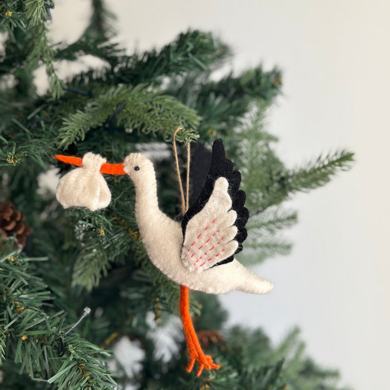A hand-felted stork ornament, a unique Christmas ornament, is nestled among the green branches of a Christmas tree. This charming white felt stork ornament has vibrant orange legs and beak. Its wings are detailed with black felt and intricate red stitching, and it carries an off-white bundle in its beak. A hemp string allows this handmade felt ornament to hang gracefully, adding a whimsical touch.