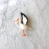 A hand-felted stork ornament is displayed on a light gray marble background. This white felt stork features a long orange beak and legs. Its wings are detailed with black and white segments, enhanced by pink stitch marks, highlighting its craftsmanship. A small white bundled felt sack is carried in its beak, symbolizing a blessing gift, with a hemp string attached for hanging.