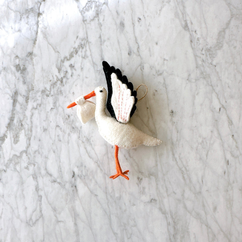 A hand-felted stork ornament is displayed on a light gray marble background. This white felt stork features a long orange beak and legs. Its wings are detailed with black and white segments, enhanced by pink stitch marks, highlighting its craftsmanship. A small white bundled felt sack is carried in its beak, symbolizing a blessing gift, with a hemp string attached for hanging.