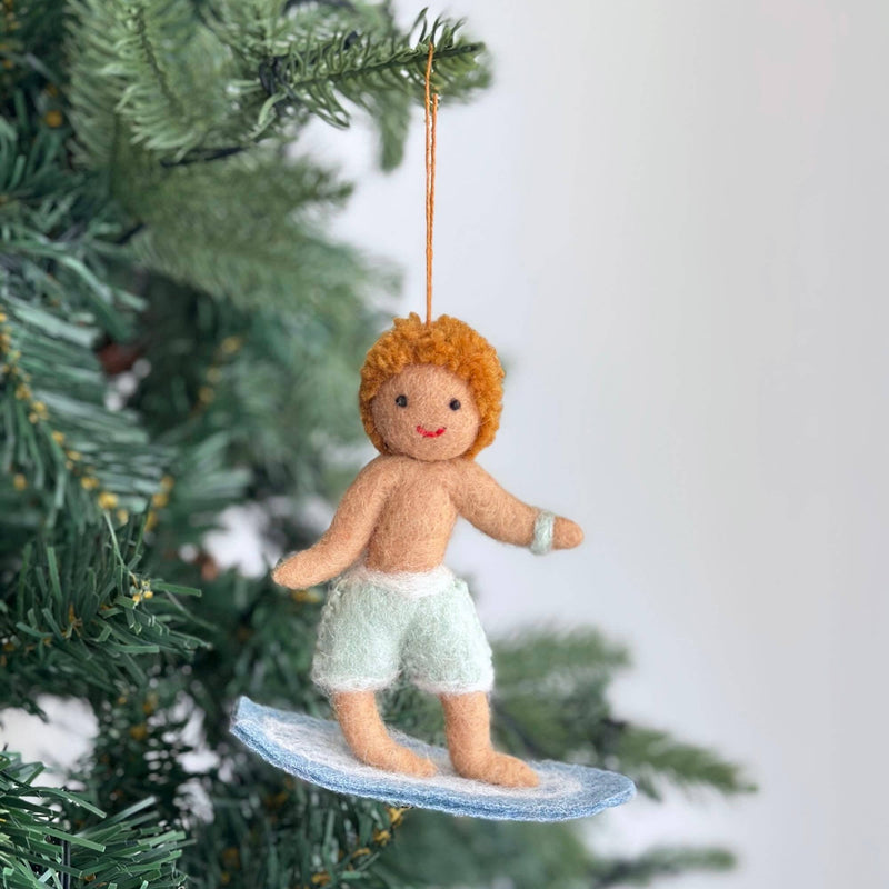 A handcrafted felt surfer boy ornament hangs on a Christmas tree, adding a joyful, festive touch. The cheerful surfer boy features orange curly hair, tan skin, and light green shorts with white trim. He rides a light blue surfboard with white detailing, ready to hang from an orange string.