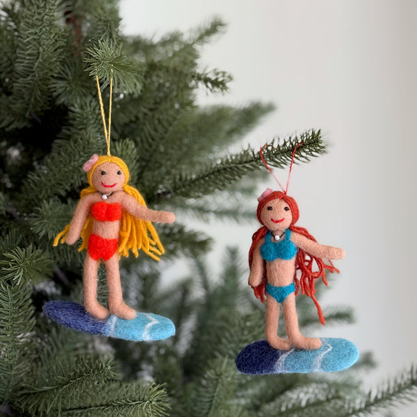 Two playful surfer girl felt ornaments hang by a jute string from a green Christmas tree branch. These handcrafted surfer ornaments feature a tan-skinned figure with bright hair, a red smile, a pink flower on her head, and a pearl choker necklace. She wears a red bikini and stands confidently on a blue and white surfboard, bringing a sense of summer fun to the holiday setting.