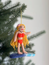 A handmade felt surfer girl ornament hangs on a Christmas tree against a soft, bright background. The tan-skinned surfer girl has long blond hair with a pink flower, wears a bright red bikini, and stands on a blue and white surfboard. A yellow string is attached for hanging.