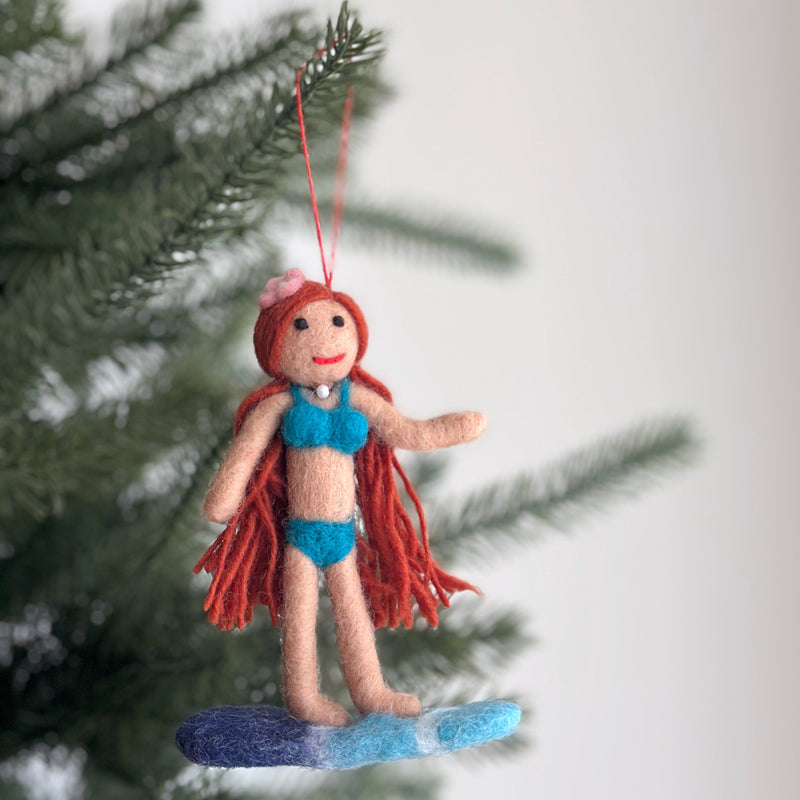 The playful felt Surfer Girl ornament hangs on a green Christmas tree, radiating a summery mood. This unique Christmas ornament features a doll figure with red hair, a flower on her head, a turquoise bikini, and a pearl choker, standing on a blue and white surfboard. A red cotton string is attached for easy hanging, bringing a whimsical touch to any holiday display.
