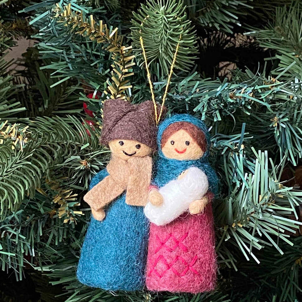 A Nativity Scene felt ornament hangs on a lush, green Christmas tree, evoking peace. This handmade felt ornament features a 3D design of the Holy Family in colorful felt. The man wears a brown hat, tan scarf, and blue robe, while the woman wears a blue shawl and pink robe with a criss-cross pattern, holding a small white figure. A gold cotton string is attached for hanging.