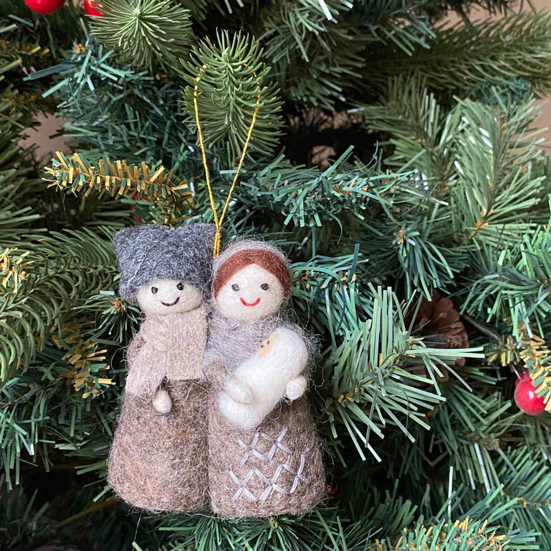 A handcrafted felt Nativity Scene ornament is suspended from a lush green Christmas tree, adorned with red berry decorations. This 3D felt ornament depicts Mary in a brown headscarf and gown, Joseph in a gray hat and robe, and baby Jesus wrapped in white, all beneath a shining star. A golden string suspends this unique Christmas ornament, adding faith and warmth to the holiday decor.