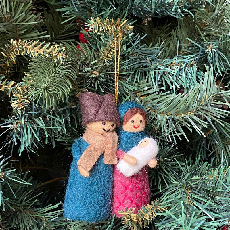 A handmade felt Nativity Scene ornament hangs delicately on a Christmas tree, suspended by a golden string. This charming 3D felt ornament depicts the Holy Family with a shining star above. The figures feature a man in a brown hat and blue robe, a woman in a blue bonnet and pink robe, and a baby in white, all with simple stitched smiles, bringing a joyful touch to this unique Christmas ornament.
