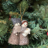 A handmade Nativity Scene felt ornament rests on a lush green Christmas tree. This 3D felt ornament depicts the Holy Family: a man in a gray hat and light brown scarf, a woman in a brown head covering, and a baby swaddled in white beneath a shining star. The figures wear brown robes with embroidered cross stitches, featuring simple stitched smiles and small white hands. A jute string is attached for easy hanging.