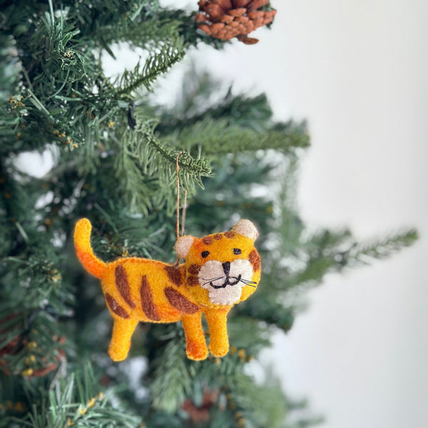 A felt tiger ornament hangs from a pine needle-covered branch of a Christmas tree, creating a joyful holiday atmosphere. This wool felt tiger ornament is yellow with brown stripes, a cream-colored belly and inner ears, and a black stitched smile with whiskers. A jute string suspends the tiger from the branch.