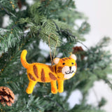 A playful wool felt tiger ornament hangs on a lush green Christmas tree, with a white wall subtly in the background. The charming felt tiger features a soft yellow body with brown stripes, perky ears, a small tail, and a sweet face with stitched whiskers. A jute string is attached for easy hanging.