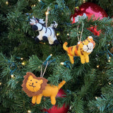 A joyful wool felt tiger ornament hangs on a vibrant green Christmas tree adorned with warm white lights and large red ornaments, creating a festive holiday scene. This handcrafted felt tiger ornament features a yellow body with orange stripes, a white smiling face, and pointed ears, suspended by a jute string.