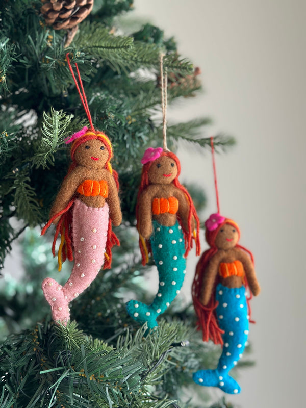 Three handmade Tropical Mermaid felt ornaments hang from the green branches of a Christmas tree. Each unique Christmas ornament features a mermaid with rich brown skin, flowing sunset-toned yarn hair, and a bright tail. The tails are stitched with pearly details and appear in pink, teal, and light blue, creating a festive holiday display.