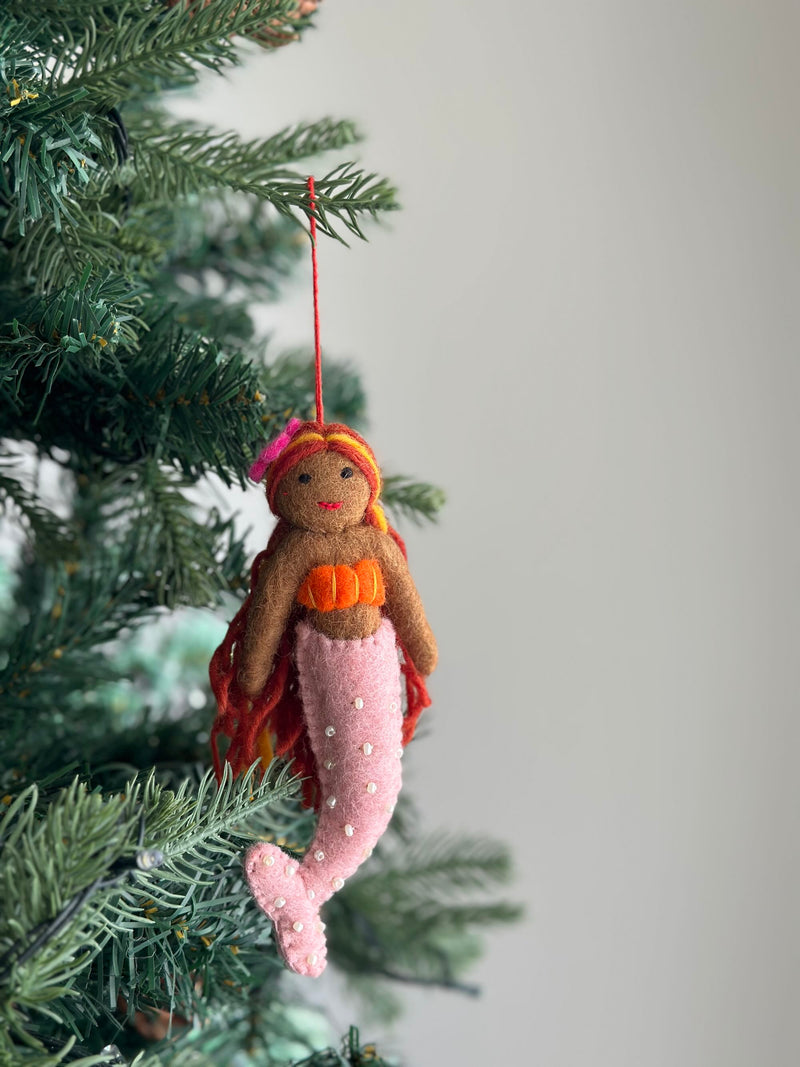 A tropical mermaid felt ornament dangles gracefully from a natural Christmas tree, set against a softly blurred light gray backdrop. This handmade felt ornament features rich brown felt skin, flowing sunset-toned yarn hair, and a bright pink tail with pearly details. A red cotton string is attached, completing its handcrafted appearance.