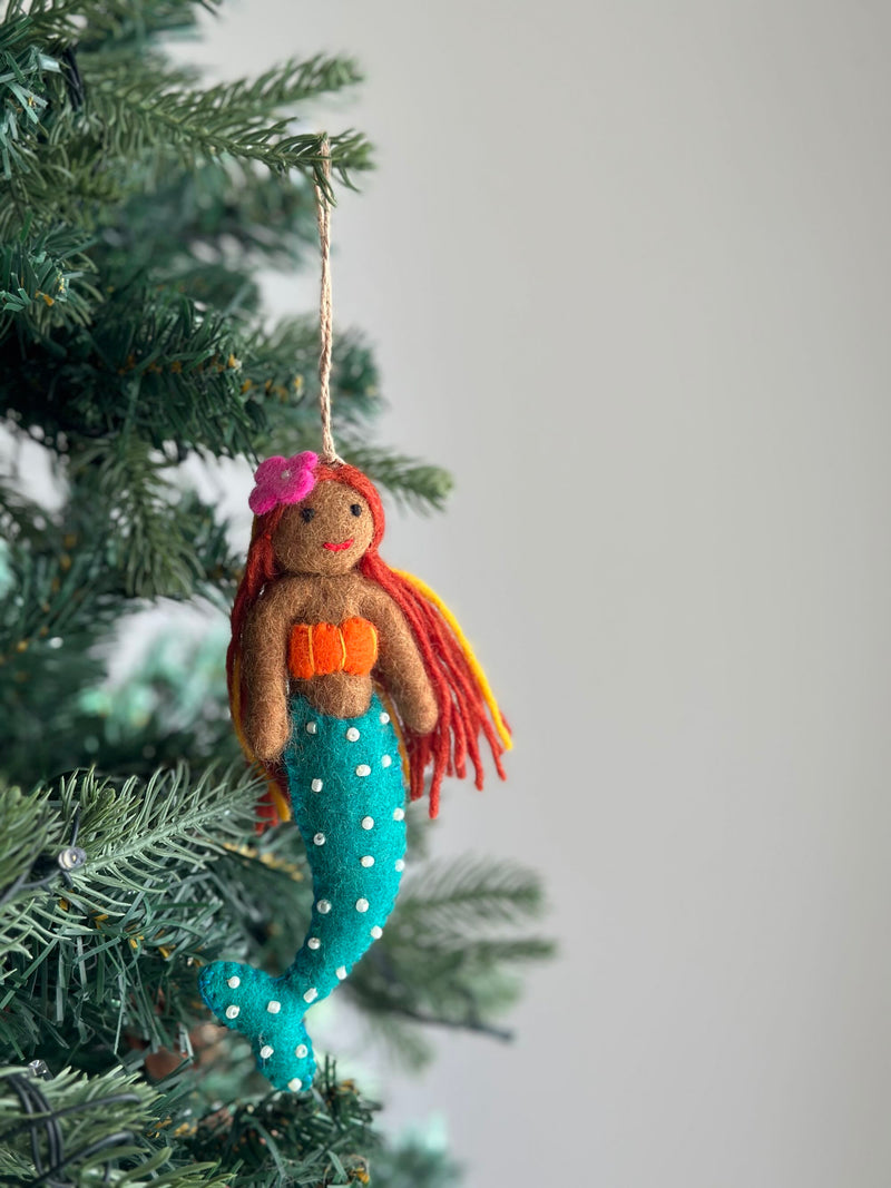 A vibrant felt tropical mermaid ornament hangs from a lush green Christmas tree, set against a soft white background. The handmade ornament features rich brown skin, flowing sunset-toned yarn hair, and a bright teal tail with pearly details. It is suspended by a jute string, embodying a handcrafted aesthetic inspired by Pacific Islander and Latina magic.