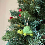 A cute felt turtle ornament hangs from the green needles of a decorated Christmas tree, adorned with red berries. This handcrafted wool felt turtle ornament is 3D, featuring a light green head and feet contrasting with a darker, textured green shell. Suspended by a jute string, this charming Christmas ornament adds a whimsical touch to the holiday decor.