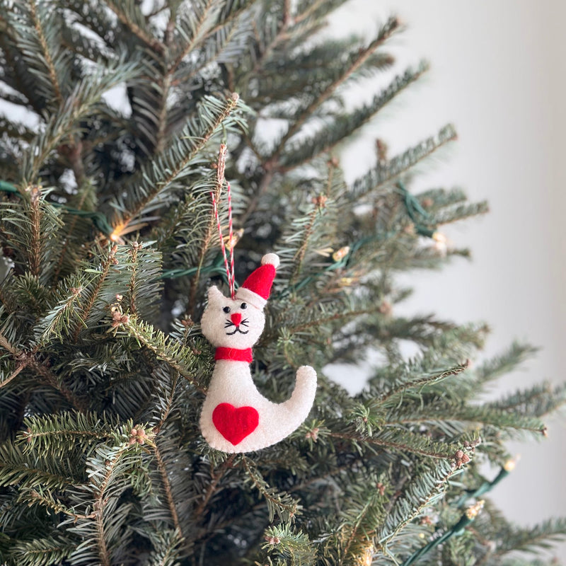 A white cat felt ornament hangs on the green branches of a Christmas tree. This charming handmade white cat wears a red Christmas hat with a white pompom, a red scarf, and has a red heart detail on its side. Its face features small black eyes, a tiny red nose, and black stitched whiskers, complete with a red and white string for hanging.