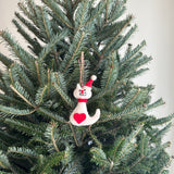 A delightful white cat felt ornament, wearing a festive red and white Santa hat, hangs by a cotton string from a green Christmas tree branch. This handmade felt ornament features a white cat with black whiskers and eyes, detailed with a red heart and a red scarf, set against a plain white wall.