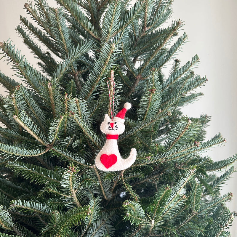 A delightful white cat felt ornament, wearing a festive red and white Santa hat, hangs by a cotton string from a green Christmas tree branch. This handmade felt ornament features a white cat with black whiskers and eyes, detailed with a red heart and a red scarf, set against a plain white wall.
