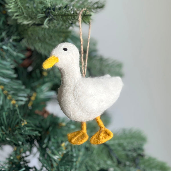A handcrafted white duck felt ornament hangs gracefully against a soft, out-of-focus Christmas tree backdrop. This cheerful white duck felt ornament is predominantly white with a small, round black eye and bright yellow bill and feet. A natural jute string is attached to the top for easy display.