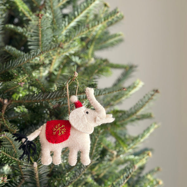 A felt white elephant ornament hangs on a vibrant Christmas tree, creating a joyful, festive atmosphere. The charming, handcrafted felt elephant has a textured, off-white body with a red Santa hat and a red blanket embroidered with a golden snowflake on its back. A black wool tail and a jute string for hanging complete the unique Christmas ornament.