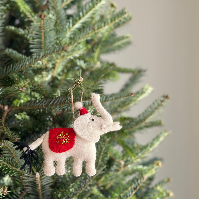 A felt white elephant ornament hangs on a vibrant Christmas tree, creating a joyful, festive atmosphere. The charming, handcrafted felt elephant has a textured, off-white body with a red Santa hat and a red blanket embroidered with a golden snowflake on its back. A black wool tail and a jute string for hanging complete the unique Christmas ornament.