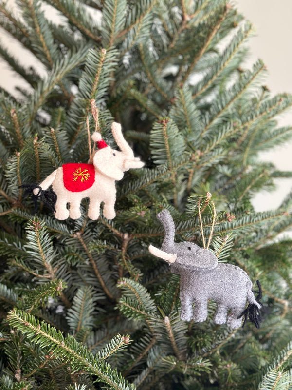 A white elephant felt ornament is nestled among the vibrant green pine needles of a Christmas tree, adding playful holiday charm. This wool felt elephant ornament features hand-stitched details, textures, and a yellow snowflake on its side. A jute string allows for easy hanging.