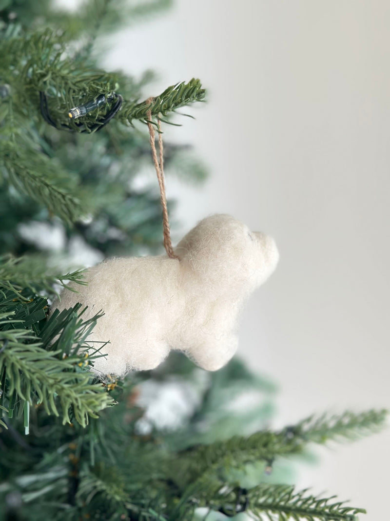 A white Maltese dog felt ornament hangs on a festive Christmas tree. This handcrafted felt ornament has a fluffy white texture, mimicking the soft fur of a Maltese dog. It hangs from a simple jute string, looped over a green pine needle branch, amidst the tree's warm glowing lights.