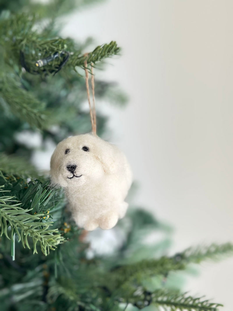 A white wool felt Maltese dog ornament hangs from a vibrant green Christmas tree branch. This handcrafted felt ornament features small black eyes, a tiny black nose, and a stitched mouth, giving it a charming, minimalist expression. A thin jute string attaches the white Maltese dog felt ornament, adding a rustic touch to the festive holiday setting.