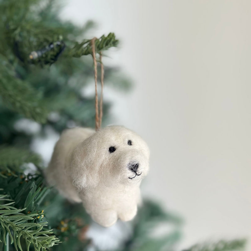 A handcrafted felt Maltese dog ornament hangs from a lush green Christmas tree branch, evoking cozy holiday warmth. This white felt Maltese dog, suspended by a jute string, features small black eyes and a stitched black nose and mouth, adding a personal and festive touch to holiday decor.