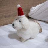 A white Maltese dog felt ornament rests on a white striped surface next to a white pillow. This charming needle-felted Christmas ornament features a fluffy white body, black eyes and nose, and a red Christmas hat with a white ball. A jute string is attached to the back for hanging.