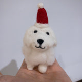 A handcrafted White Maltese Dog felt ornament is gently held in a female hand, positioned against a plain white wall. This unique felt ornament features fluffy white wool felt with small, round black eyes and a stitched black nose and mouth, giving it a sweet expression. A small, bright red felted Christmas hat with a tiny white pompom adorns its head, adding a festive touch to this Christmas ornament.