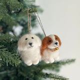 A white Maltese dog felt ornament hangs joyfully from a lush green Christmas tree branch, suspended by a jute string. This charming wool felt ornament features a round, fluffy body, small black eyes, and a stitched nose, embodying a sweet, expressive face. Just beside it, a regal Cavalier King Charles Spaniel felt ornament dangles gracefully, its soft chestnut and white colors complementing the Maltese. Both ornaments bring a cozy, festive charm to the scene, set against a blurred white wall.