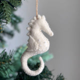 A white seahorse felt ornament hangs on a subtly lit Christmas tree branch. This handcrafted felt ornament features a textured white felt surface embellished with small, clear beads. A jute string is attached, completing its decorative charm for a classic Christmas setting.