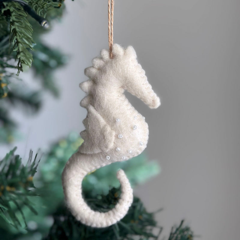 A white seahorse felt ornament hangs on a subtly lit Christmas tree branch. This handcrafted felt ornament features a textured white felt surface embellished with small, clear beads. A jute string is attached, completing its decorative charm for a classic Christmas setting.