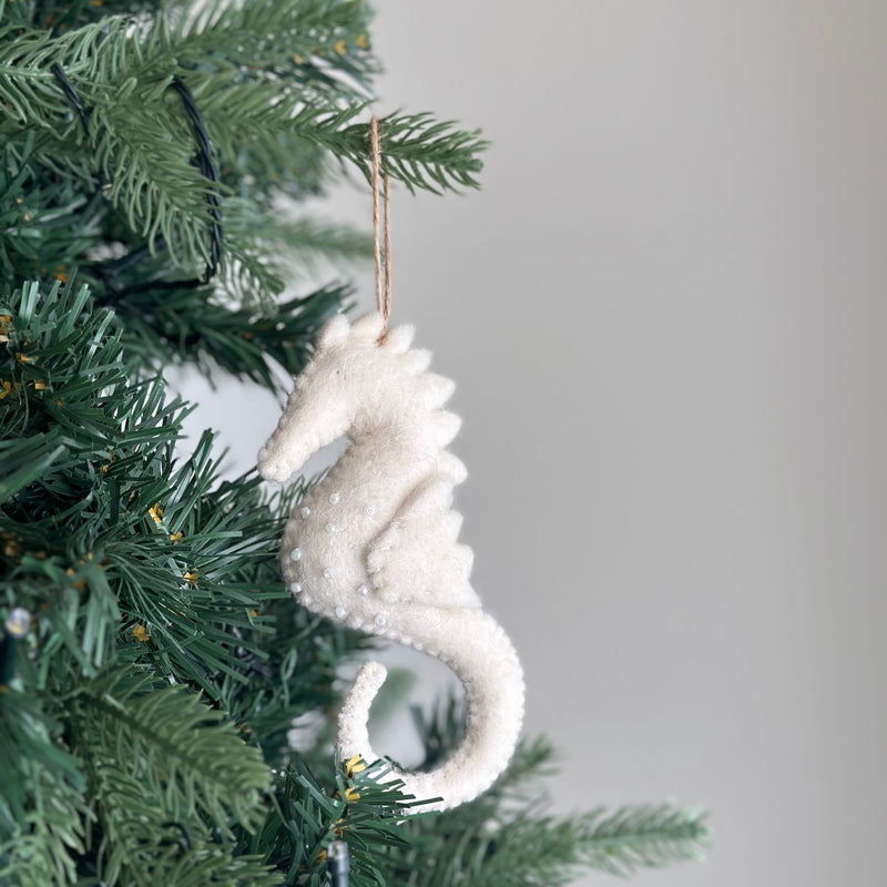 A white seahorse felt ornament hangs gracefully from a jute string on a deep green Christmas tree, creating a serene festive scene. This handcrafted felt ornament is shaped from soft white felt, detailed with delicate white bead accents, a curved tail, and subtle spiky details along its back.