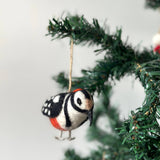A needle felted woodpecker ornament hangs from a vibrant Christmas tree branch, suspended by a jute string. The handcrafted woodpecker felt ornament has a predominantly white body with black stripes, a long black beak, and a distinct red patch on its head, adding a cheerful, natural holiday charm.