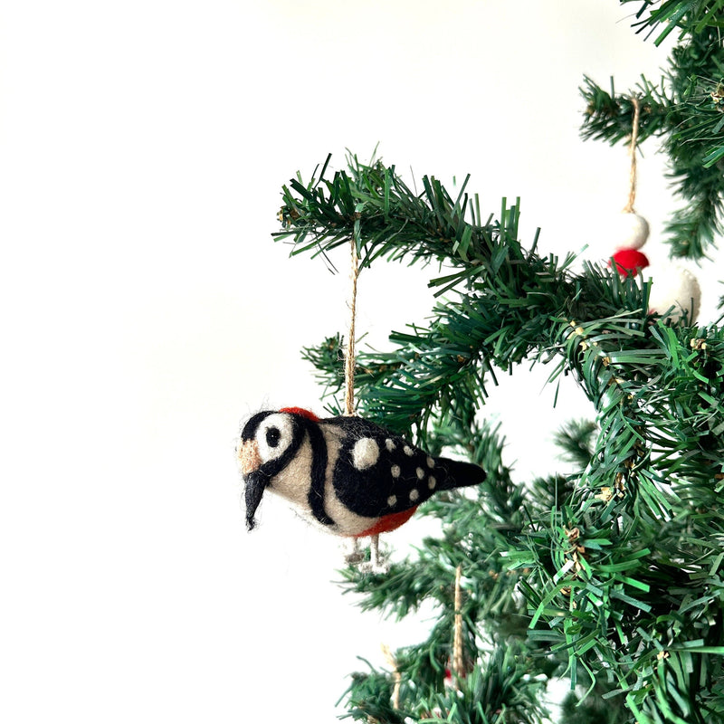 A felt woodpecker ornament hangs from a branch of a lush green Christmas tree, adding rustic charm to the festive setting. This handcrafted felt ornament is detailed in white, black, and red felt, showcasing realistic plumage and a distinctive beak. It is suspended by a jute string.