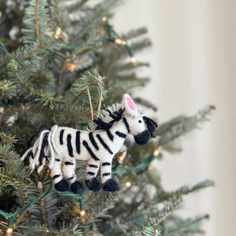 A needle-felted zebra ornament hangs on a Christmas tree adorned with soft, warm fairy lights. This approximately 4-inch tall wool felt zebra ornament features intricate needle-detailed black and white stripes, a fluffy mane, and pink inner ears. A black yarn tail with white stripes hangs below, as the unique Christmas ornament is suspended by a jute string from a green branch.