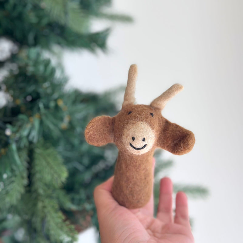 A handcrafted wool felt ox or cow finger puppet is gently held by a female hand, set against a soft-focus Christmas pine tree backdrop. The light brown puppet features floppy ears, distinctive curved horns, and a cheerful stitched smile, showcasing its charming details and artisanal quality.