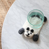 A single handmade wool felt panda coaster, ethically crafted by Nepalese female artisans, rests flat on a round marble table beneath a clear glass of water. This heat-resistant felt panda coaster is primarily white with black accents forming the panda's ears and distinctive markings, detailed with small stitched eyes and a smile. A natural woven rug peeks from the cozy background.
