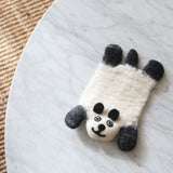 A handcrafted wool felt panda coaster rests on a white marble table next to a woven rug. This felt panda coaster features a cream body with black ears, paws, and facial details like round eyes and a smiling mouth. Crafted from soft sheep wool felt, this unique coaster offers a playful yet practical touch to home decor.