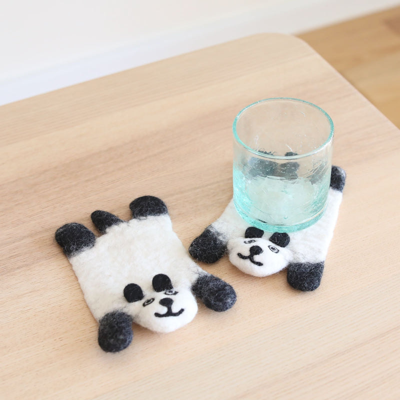 Two felt panda coasters, part of a set, rest on a light wooden table against a white wall. These playful, handcrafted wool felt panda coasters are white with black ears, arms, and facial features. Each ethically made, heat-resistant coaster measures approximately 4 inches in diameter. One panda coaster is displayed full-body, while another peeks out from under a glass of water.